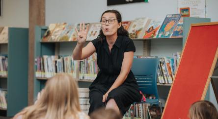 A woman in a black shirt sits while telling a story, holding up four fingers, in front of a group of attentive children. A empty red felt board sits to her right.