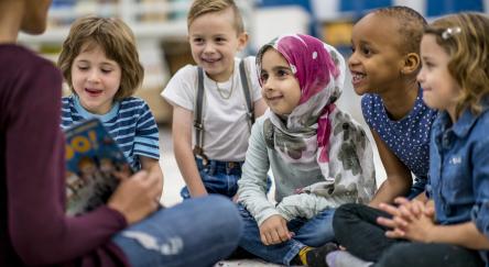 A diverse group of four children sitting on the floor. An adult reads happily to them in a cozy setting.