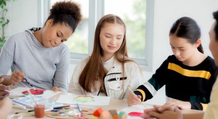 A group of 3 diverse teen girls sitting around a table colouring, drawing, and painting.