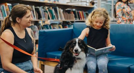 A young girl sits on a blue couch petting and reading to a black and white dog. The dog an a caregiver sit on the floor next to the reading child.
