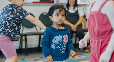 Three toddlers try to catch bubbles during Toddler Social, while a parent watches in the background.