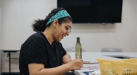 South Asian woman (30s) with teal headband sits at table surrounded by art supplies while she colours a sheet.