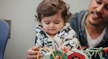 A smiling dad and a baby girl engage in playtime, surrounded by a variety of fun toys at the library.