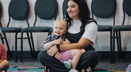 A Caucasian mom (30s) sits on floor with toddler girl in her lap at interactive library program.