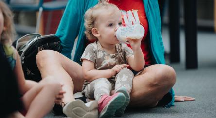 A mom and baby girl sit on the library floor together listening to stories while the baby drinks from a bottle.