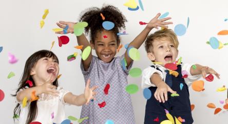A multi-ethnic group of three small kids, throw colourful confetti into the air while laughing.