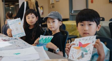 Asian boy (8) and two Asian girls (8) hold up their painted pictures.