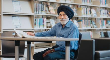 A senior South Asian man (70s) looking up sits at table with a book at the library.