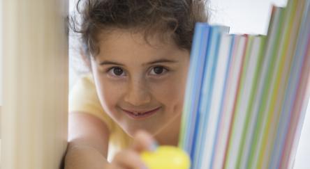 A young female child peeks through a bookshelf to grab a yellow Easter egg.