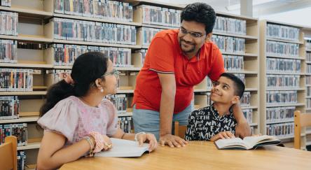 A South Asian girl (14) and South Asian boy (10) study at a library table while their South Asian dad (40s) leans over smiling proudly.