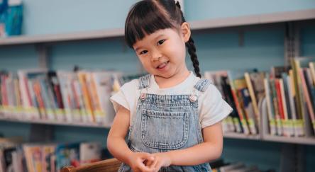 Young Asian girl (5) with braids does crafts at a table with the library book shelves in the background.