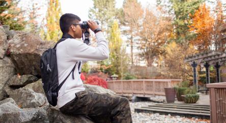 South asian teen boy (18) uses with binoculars and backpack looks at birds in the distance.