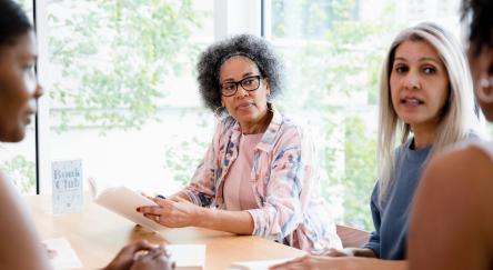 The multiracial group of women listen to the unrecognizable leader at the head of the table.