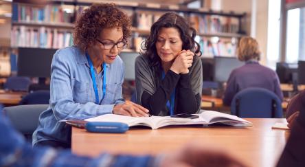 Two women sit together at table in library discussing and working on their writing skills. Library stacks, and public computers are used by other customers in the background.