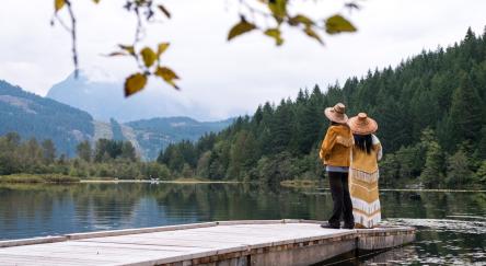 Two Indigenous people stand at the end of a dock looking out at the lake.