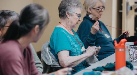 Four older ladies sit in a row at a table. They are all working on different knitting and crocheting projects and enjoying each other's company.