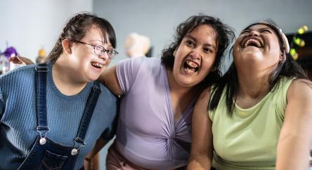 Three young female adults with diverse abilities embrace and laugh with each other.