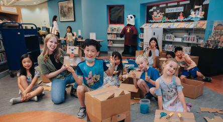 A large group of children and a female library staffer, cut up cardboard using Makedo tools and screws to create imaginative cardboard creations like a dragon head.