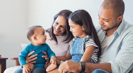 Multiethnic family playing with happy baby son at home. Parent and children relaxing together on the sofa at home in the living room. Little girl sitting on leg of dad looking her new cute brother.