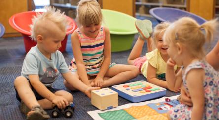 A group of children engaged in play, learning to code with Cubetto through stories and hands-on play, in a bright and inviting library space.