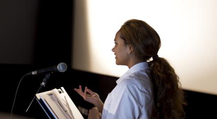 A female (20s) stands in front of a microphone, reading a poem to the unseen audience in front of her.