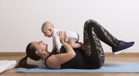 A mom, working out laying on her back, holds her infant above her. She bonds with her infant while doing physical activity.