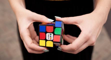 A pair of hands at waist level, with white painted nails, work to solve a Puzzle Cube.