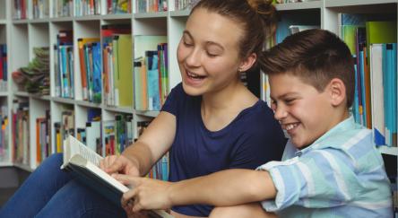 A teen girl (15) and boy (8) practice reading together in library.