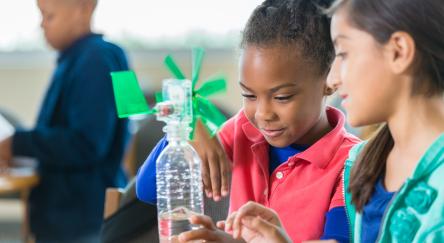 African American and Hispanic elementary age little girls are building a windmill from a water bottle during an after school science program in a public library. Students are learning science, technology, engineering, and math while working on a project together.
