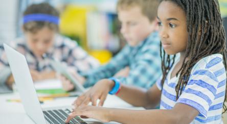 A multi-ethnic group of children are indoors in a library program. They are wearing casual clothing. They are working together on a laptop computer learning to code together.
