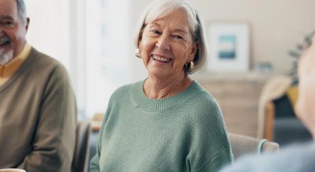 A group of seniors gather to socialize. A senior woman looks into the camera and smiles, while a man appears in the side of the image.