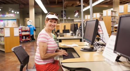 A older woman  in shorts, t-shirt, and hat, sits at a library computer in a library. She looks at the camera while typing. Bookshelves can be seen in the background.