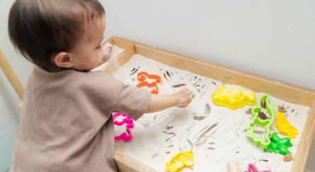 Toddler playing kinetic sand with plastic shapes on sensory table.