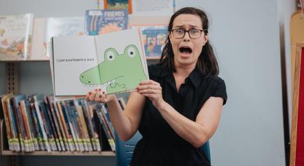 A librarian reads a storybook to a group of kids. Her face makes an animated scared expression as she shows the kids the image of a crocodile in the book.