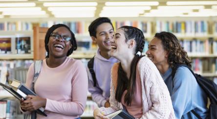 A multiracial group of four high school students carrying backpacks, laptops and textbooks, walking in a library, conversing, laughing. They are teenagers, 15 and 16 years old.