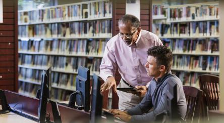Two multi-ethnic men in the library looking at a computer screen and conversing. A mature man in his 40s is sitting at the desktop PC and a senior African-American man in his 60s is standing beside him.