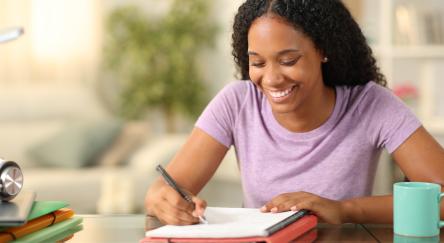 Teen girl, sitting at a counter writing on paper that is stacked on top of a binder. A coffee mug and other desktop items can be seen in the surrounding area.