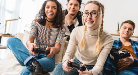 A enthusiastic multi-ethnic group of two teen girls, and two teen boys holding video game controllers.