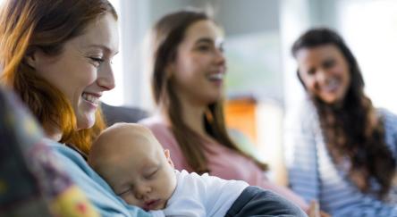 Smiling mother holding sleeping newborn son sitting by friends at a mom's group. 
