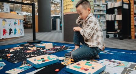 Young boy plays with Cubetto, a friendly wooden robot that helps teach coding, on the library floor.