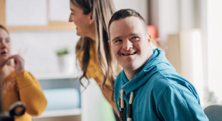 A young Caucasian down syndrome man (20s) and women in the background gather in a social setting.