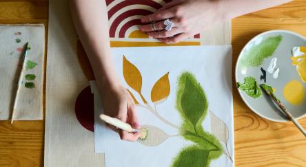 Above angle of hands of young female artist with paintbrush painting green leaves on paper while sitting by table in workshop.