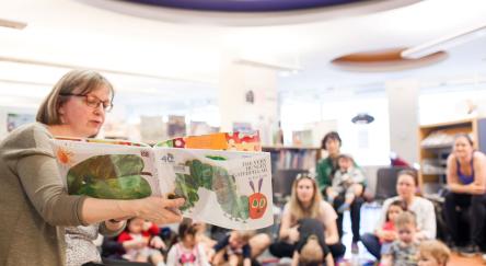 Female library staffer (50s) reads 'The Very Hungry Caterpillar' to a large audience of kids and parents. Audience sits on a blue carpet on the library's floor. 