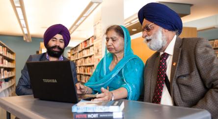A South Asian man with purple turban assists a South Asian woman wearing a teal langa and her South Asian man in blue turban with a laptop.