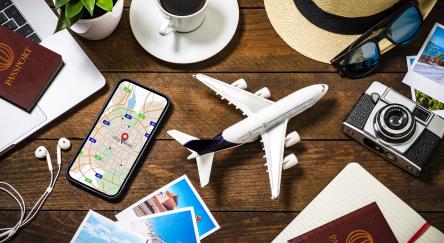 Top view of an tiny airplane surrounded by various traveling stuff such as a coffee cup, a camera with some snapshots, a smartphone with a map on the screen, an opened notebook, two passports with boarding passes and some summer accessories like a hat and sunglasses. All the objects are on a rustic wooden desk.