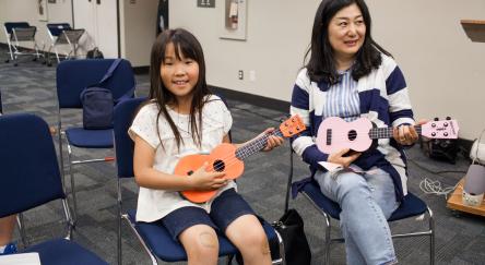 A mom (40s) and daughter (9) practice playing ukuleles together in library program room.
