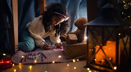 Little girl writing down letter under the table surrounded by fairy lights spread out over the floor.
