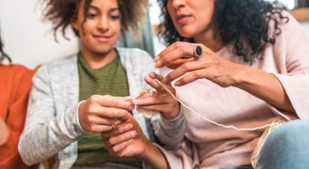 An older woman teaches a teenage girl basic knitting stitches in a relaxed, homey environment.
