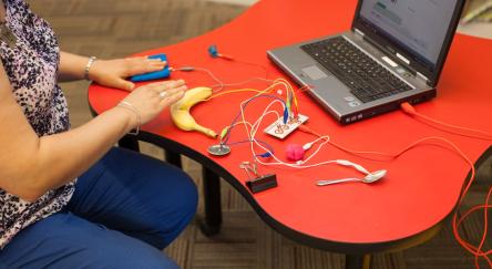 A person plays with a Makey Makey circuit board device by connecting it to various items to create computer commands.