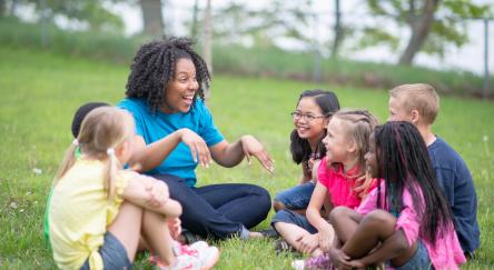 A female librarian with a group of children in the park. She is telling them a story. The children are happy and smiling.